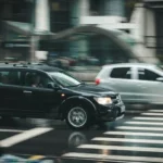 A black SUV and a silver hatchback drive past each other on a wet city street, blurred by motion, with a pedestrian crosswalk in the foreground—capturing a tense moment that hints at the potential for an accident 