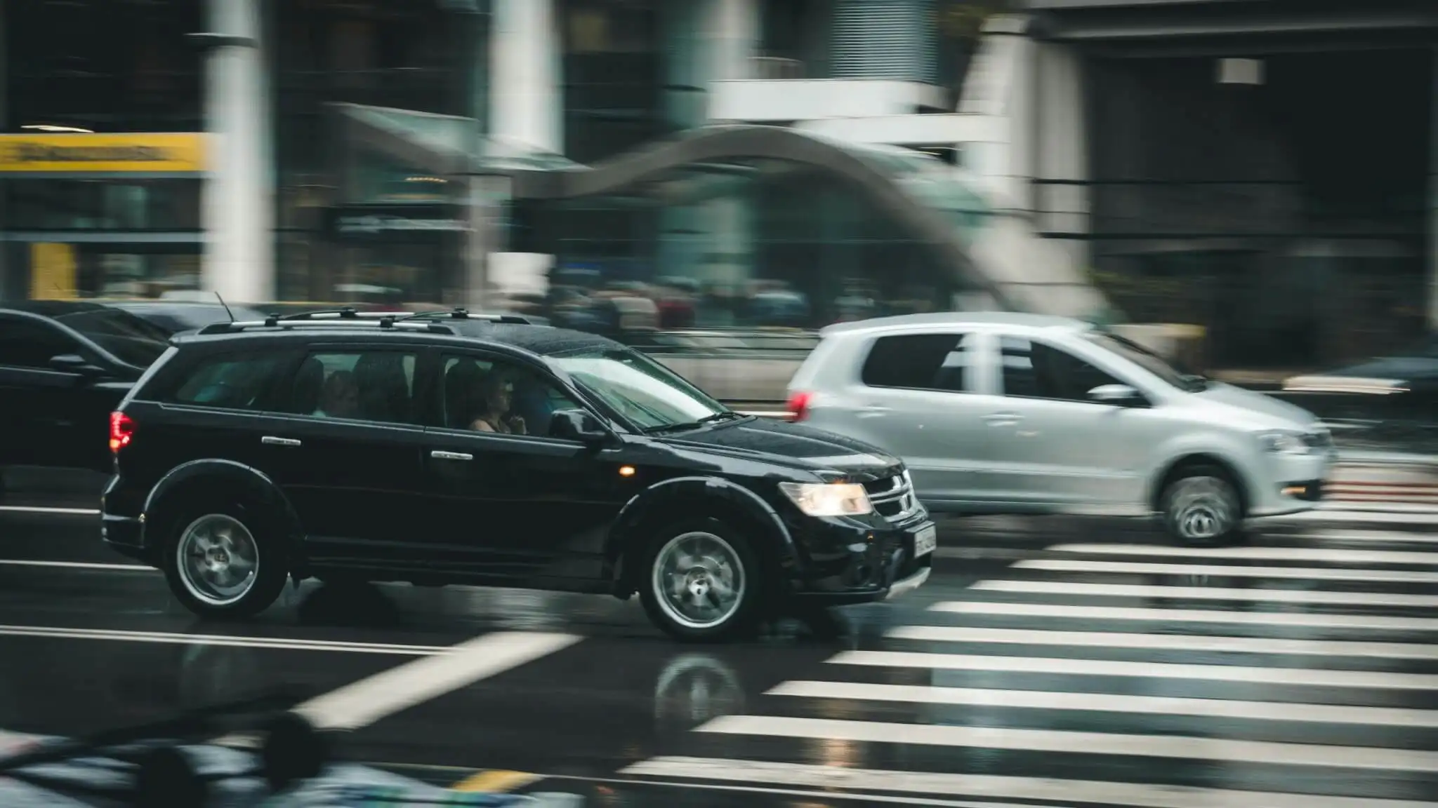 A black SUV and a silver hatchback drive past each other on a wet city street, blurred by motion, with a pedestrian crosswalk in the foreground—capturing a tense moment that hints at the potential for an accident 