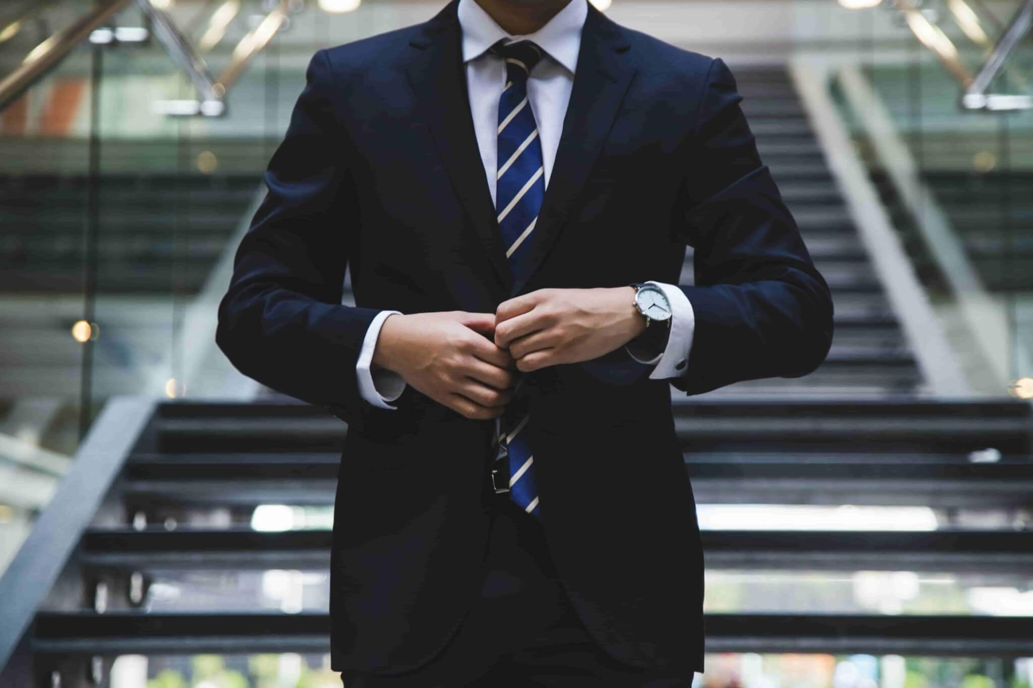 A person in a dark suit and striped tie adjusts their jacket while standing in front of a staircase in a modern building, perhaps pondering, "Do I Need a Personal Injury Lawyer?. - Ghassemi Law Group A person in a dark suit and striped tie adjusts their jacket while standing in front of a staircase in a modern building, perhaps pondering, "Do I Need a Personal Injury Lawyer?.