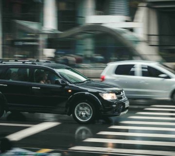 A black SUV and a silver hatchback drive through a city crosswalk on a rainy day, motion blur suggesting movement—raising questions like, How Much Does a Car Accident Lawyer Cost in Irvine?. - Ghassemi Law Group A black SUV and a silver hatchback drive through a city crosswalk on a rainy day, motion blur suggesting movement—raising questions like, How Much Does a Car Accident Lawyer Cost in Irvine?.