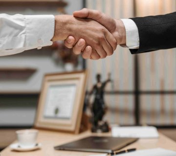 Two people in business attire shake hands across a desk, with a certificate, statuette, and coffee cup in the background—setting the scene to discuss topics like "How Much Does a Personal Injury Lawyer Cost in Irvine?. - Ghassemi Law Group Two people in business attire shake hands across a desk, with a certificate, statuette, and coffee cup in the background—setting the scene to discuss topics like "How Much Does a Personal Injury Lawyer Cost in Irvine?.