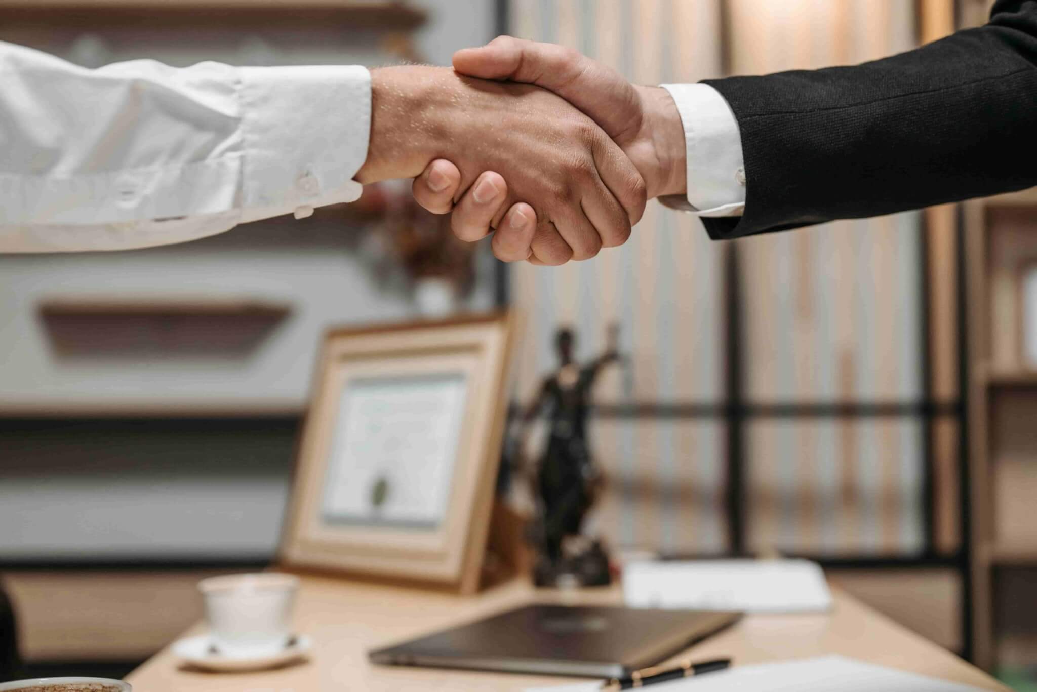 Two people in business attire shake hands across a desk, with a certificate, statuette, and coffee cup in the background—setting the scene to discuss topics like "How Much Does a Personal Injury Lawyer Cost in Irvine?. - Ghassemi Law Group Two people in business attire shake hands across a desk, with a certificate, statuette, and coffee cup in the background—setting the scene to discuss topics like "How Much Does a Personal Injury Lawyer Cost in Irvine?.