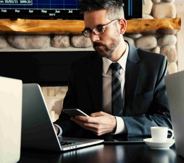 A man in a suit sits at a desk using a laptop and holding a smartphone, researching How to Find the Best Personal Injury Lawyer in Irvine, with two laptops, a coffee cup, and a stone fireplace in the background. - Ghassemi Law Group A man in a suit sits at a desk using a laptop and holding a smartphone, researching How to Find the Best Personal Injury Lawyer in Irvine, with two laptops, a coffee cup, and a stone fireplace in the background.