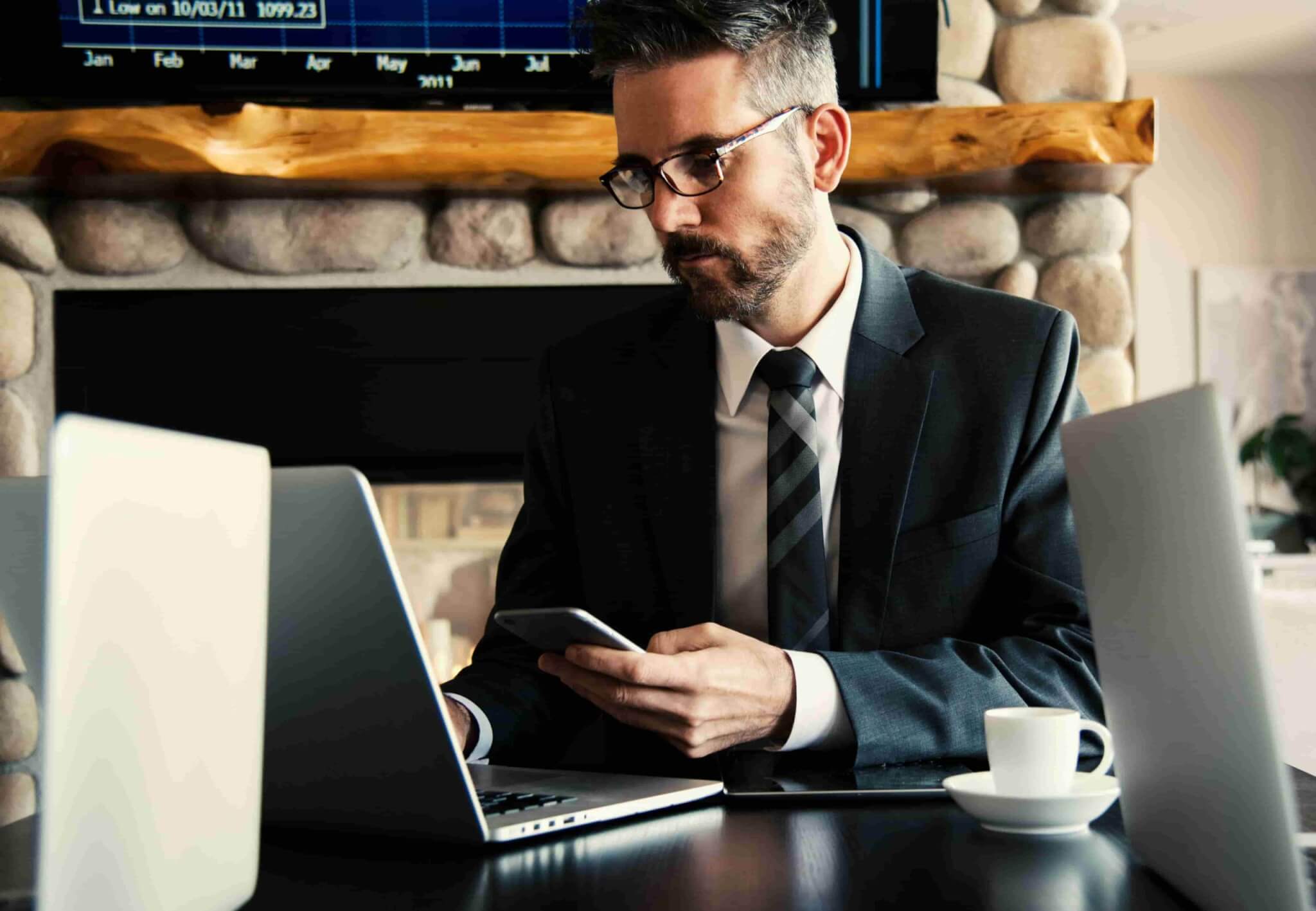 A man in a suit sits at a desk using a laptop and holding a smartphone, researching How to Find the Best Personal Injury Lawyer in Irvine, with two laptops, a coffee cup, and a stone fireplace in the background. - Ghassemi Law Group A man in a suit sits at a desk using a laptop and holding a smartphone, researching How to Find the Best Personal Injury Lawyer in Irvine, with two laptops, a coffee cup, and a stone fireplace in the background.