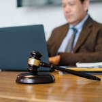 A wooden gavel rests on a desk in the foreground, while a lawyer for a minor car accident works on a laptop in the blurred background. - Ghassemi Law Group A wooden gavel rests on a desk in the foreground, while a lawyer for a minor car accident works on a laptop in the blurred background