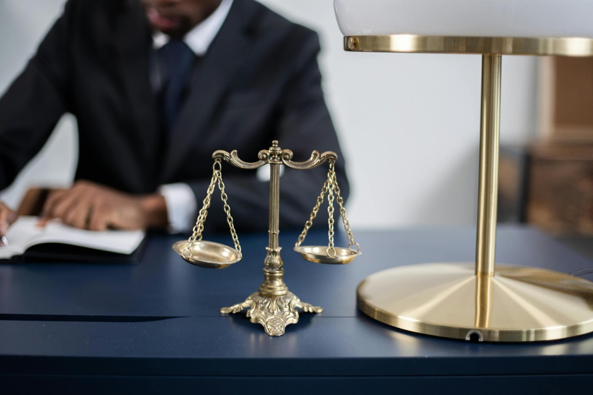 A brass balance scale sits on a desk next to a lamp, while a car accident lawyer in Irvine writes in a notebook in the background. - Ghassemi Law Group A brass balance scale sits on a desk next to a lamp, while a car accident lawyer in Irvine writes in a notebook in the background.