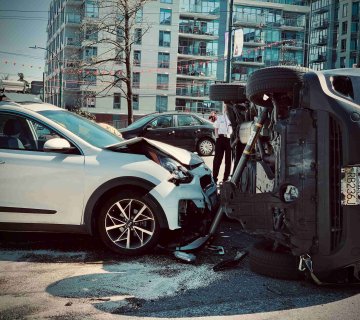 A white SUV with a damaged front end is stopped next to a black vehicle that has rolled onto its side in an urban area, as bystanders look on—an incident where contacting a personal injury lawyer may be necessary. - Ghassemi Law Group A white SUV with a damaged front end is stopped next to a black vehicle that has rolled onto its side in an urban area, as bystanders look on—an incident where contacting a personal injury lawyer may be necessary.