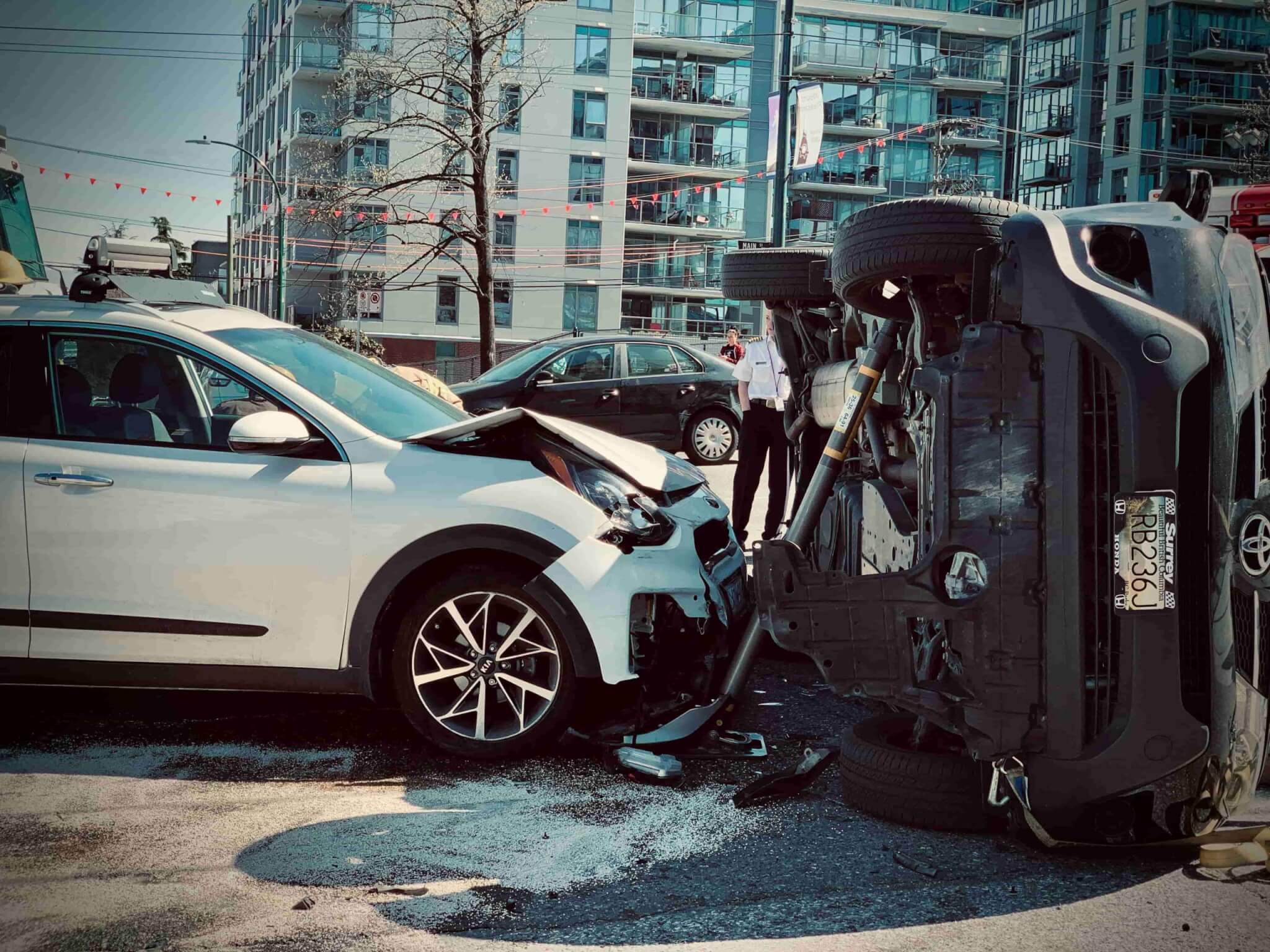 A white SUV with a damaged front end is stopped next to a black vehicle that has rolled onto its side in an urban area, as bystanders look on—an incident where contacting a personal injury lawyer may be necessary. - Ghassemi Law Group A white SUV with a damaged front end is stopped next to a black vehicle that has rolled onto its side in an urban area, as bystanders look on—an incident where contacting a personal injury lawyer may be necessary.