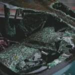 A car windshield is severely shattered, with large broken glass pieces scattered on the car’s hood and interior, suggesting the possibility of a burn injury amid the chaotic aftermath 