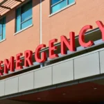 Red “EMERGENCY” sign on the exterior of a hospital building, often indicating the critical care entrance for accident victims, with brick walls and glass windows visible in the background 