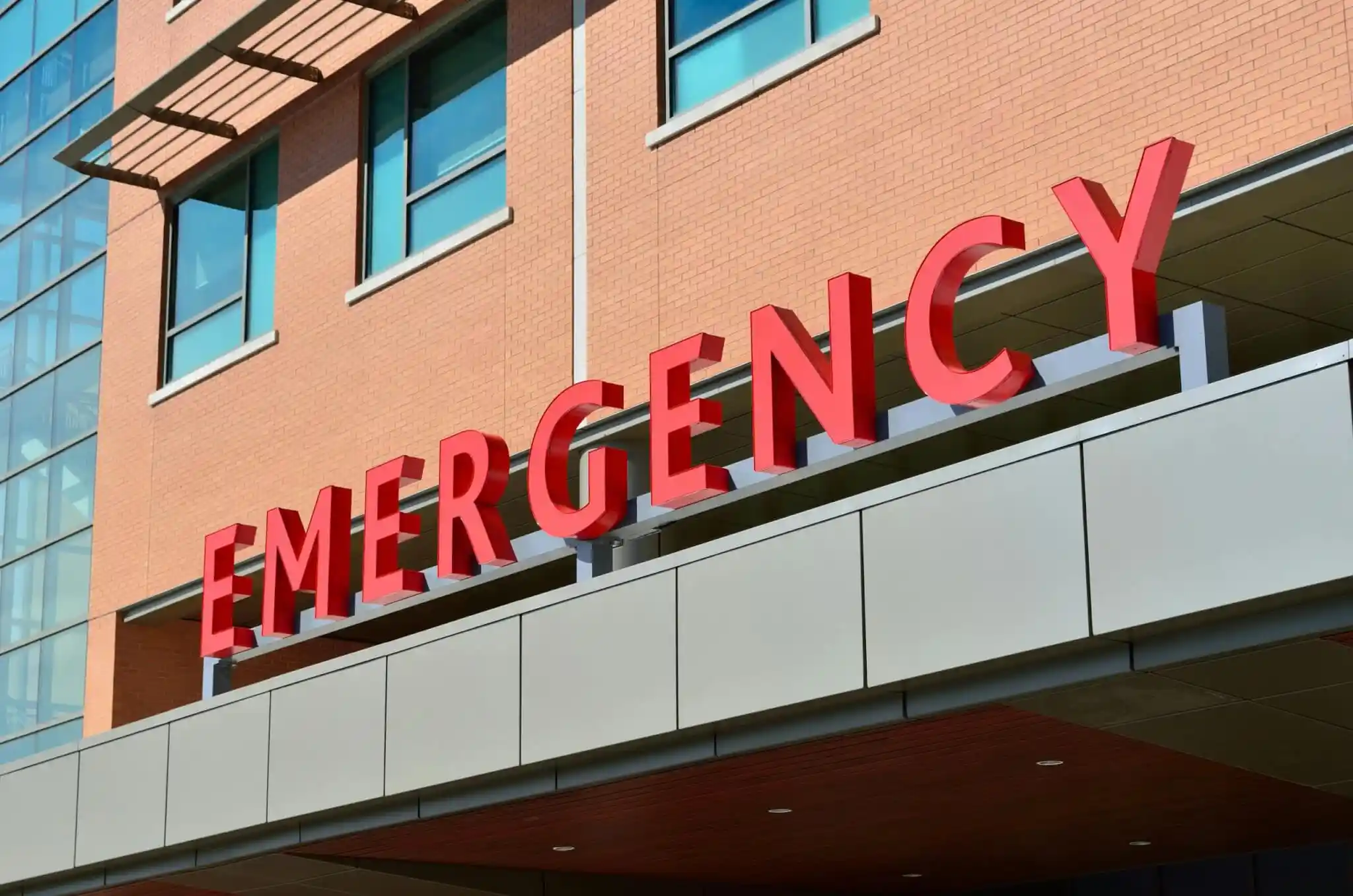 Red “EMERGENCY” sign on the exterior of a hospital building, often indicating the critical care entrance for accident victims, with brick walls and glass windows visible in the background 