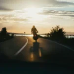 A person rides a motorcycle down an empty road at sunset, with trees on both sides and water visible in the background 