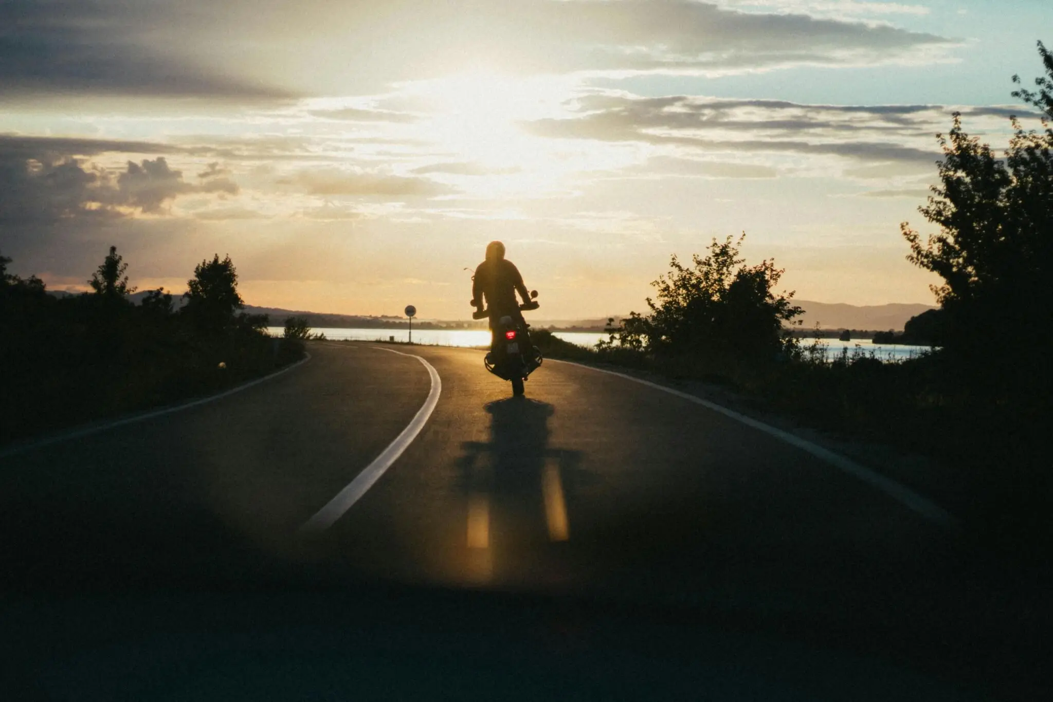 A person rides a motorcycle down an empty road at sunset, with trees on both sides and water visible in the background. - Ghassemi Law Group A person rides a motorcycle down an empty road at sunset, with trees on both sides and water visible in the background