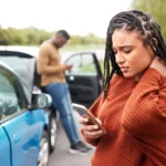 Two people stand by their cars after a crash  A woman in the foreground looks at her phone, holding her neck, while a man in the background uses his phone beside an open car door 