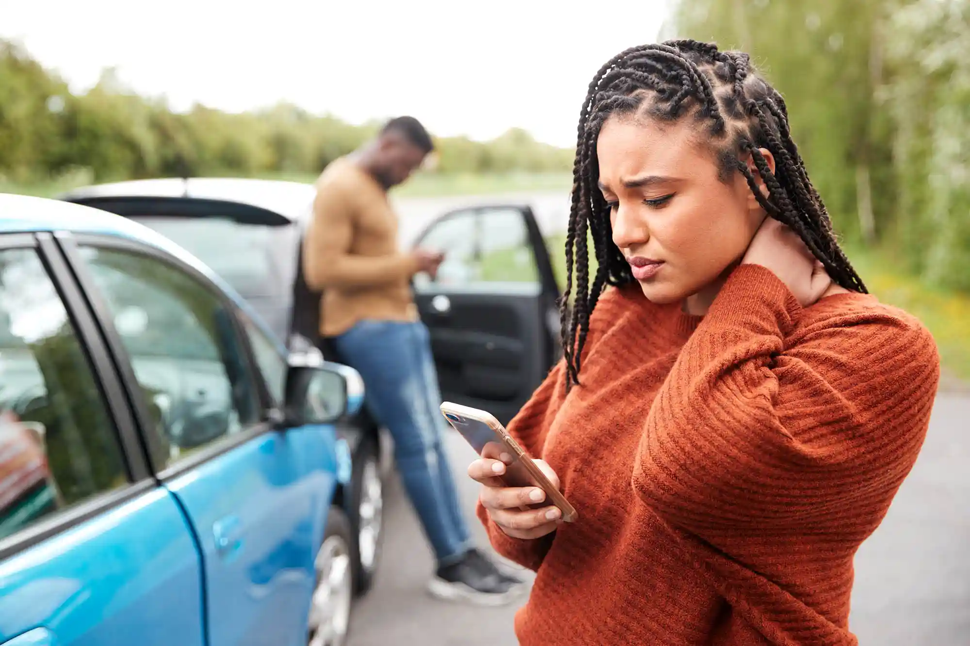 Two people stand by their cars after a crash  A woman in the foreground looks at her phone, holding her neck, while a man in the background uses his phone beside an open car door 