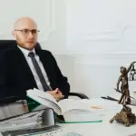 A man in a suit sits at a desk with legal books, documents, and a small statue of Lady Justice—ready to advise you on questions like "How Much Does a Car Accident Lawyer Cost in Irvine" in an office setting