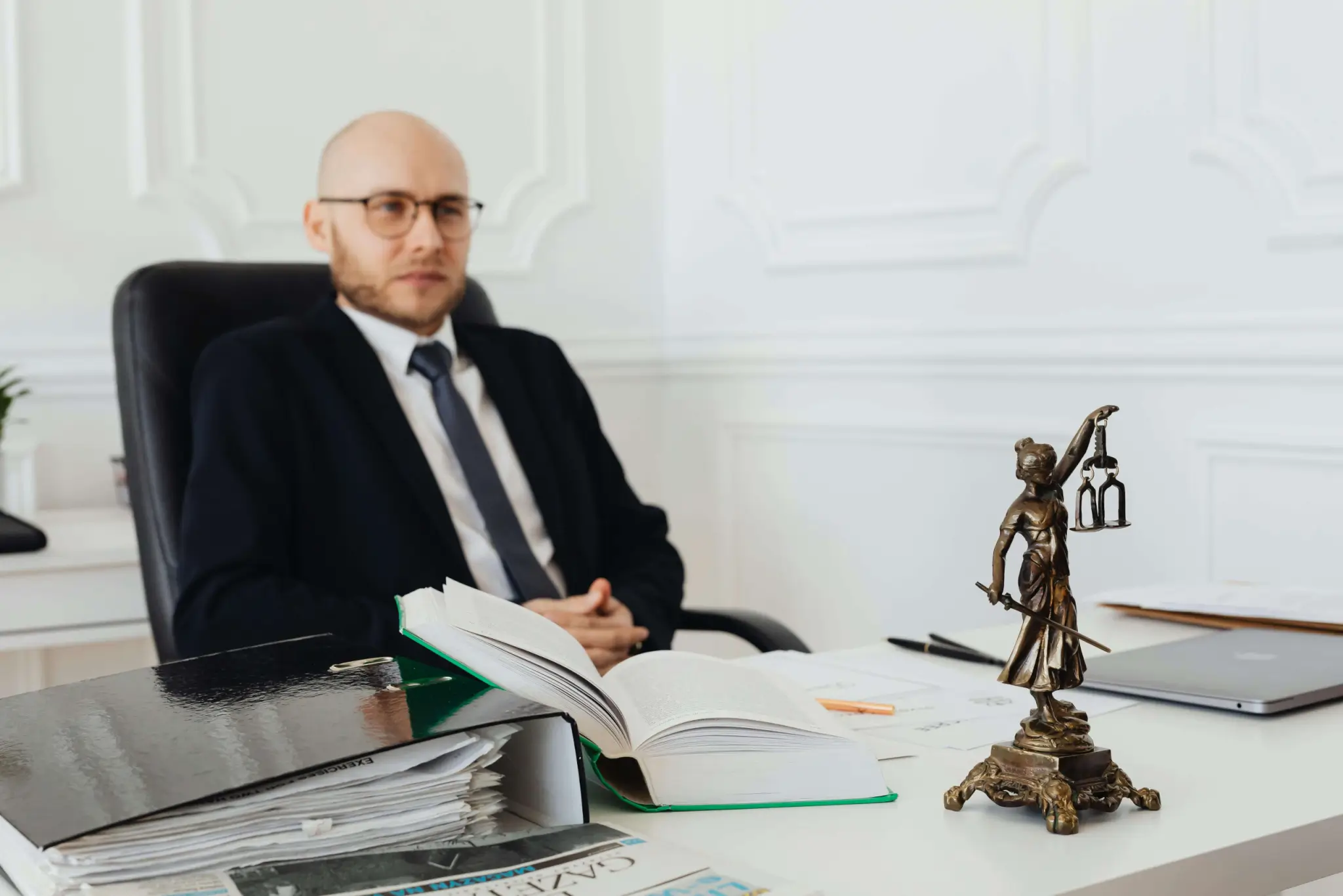 A man in a suit sits at a desk with legal books, documents, and a small statue of Lady Justice—ready to advise you on questions like "How Much Does a Car Accident Lawyer Cost in Irvine" in an office setting. - Ghassemi Law Group A man in a suit sits at a desk with legal books, documents, and a small statue of Lady Justice—ready to advise you on questions like "How Much Does a Car Accident Lawyer Cost in Irvine" in an office setting