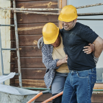 Two construction workers wearing yellow helmets; one is helping the other walk, possibly injured from a workplace slip and fall, in front of a wooden building. - Ghassemi Law Group Two construction workers wearing yellow helmets; one is helping the other walk, possibly injured from a workplace slip and fall, in front of a wooden building