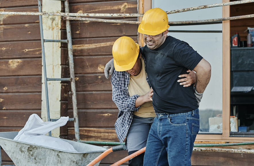 Two construction workers wearing yellow helmets; one is helping the other walk, possibly injured from a workplace slip and fall, in front of a wooden building. - Ghassemi Law Group Two construction workers wearing yellow helmets; one is helping the other walk, possibly injured from a workplace slip and fall, in front of a wooden building