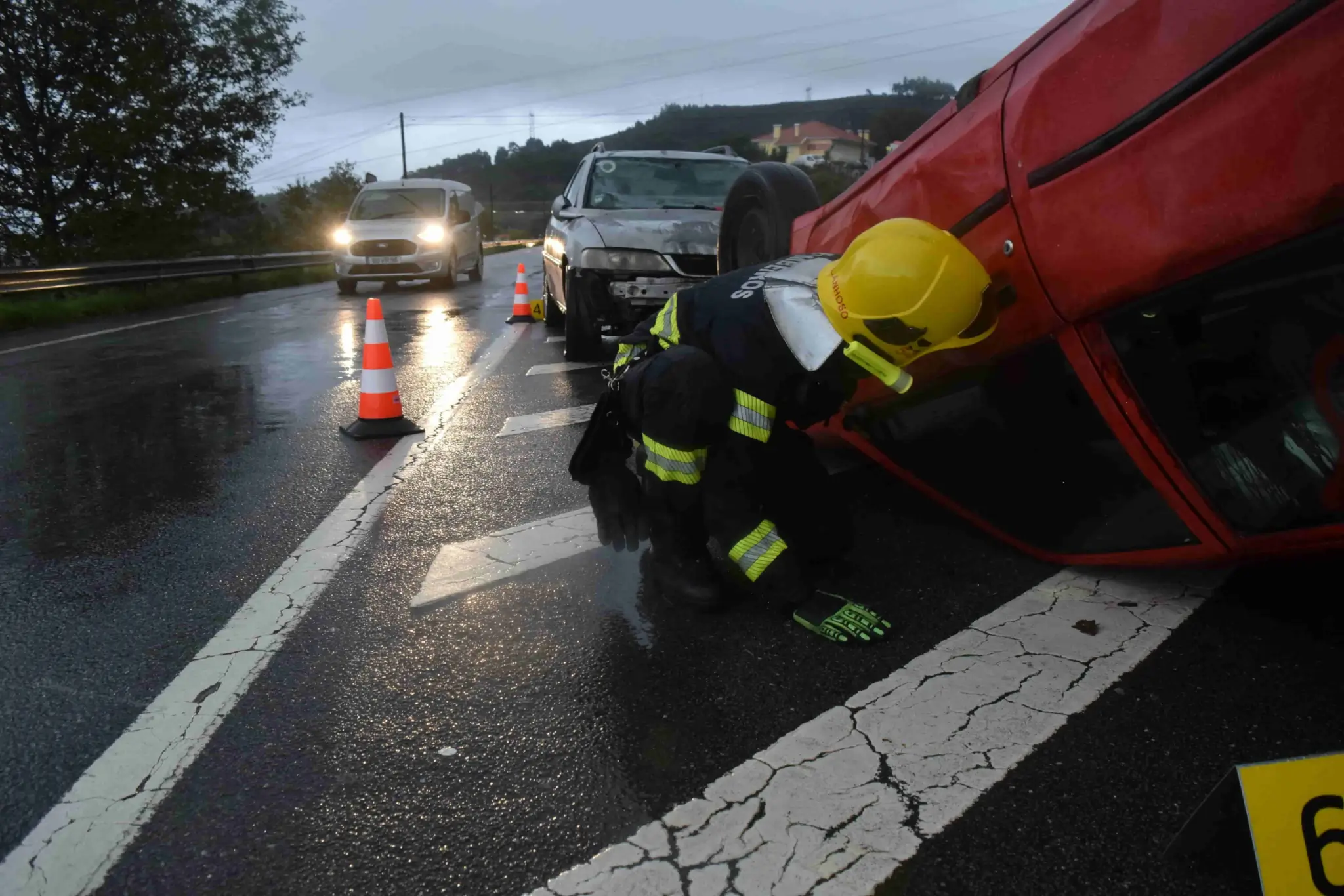 A firefighter examines an overturned red car after an accident on a wet road, with cones and another damaged vehicle nearby—raising the question: What Should I Do Immediately After a Car Accident in Irvine?. - Ghassemi Law Group A firefighter examines an overturned red car after an accident on a wet road, with cones and another damaged vehicle nearby—raising the question: What Should I Do Immediately After a Car Accident in Irvine?
