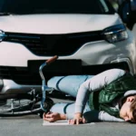 A person lies on the ground next to a fallen bicycle in front of a stopped white car, suggesting a collision at a crosswalk 