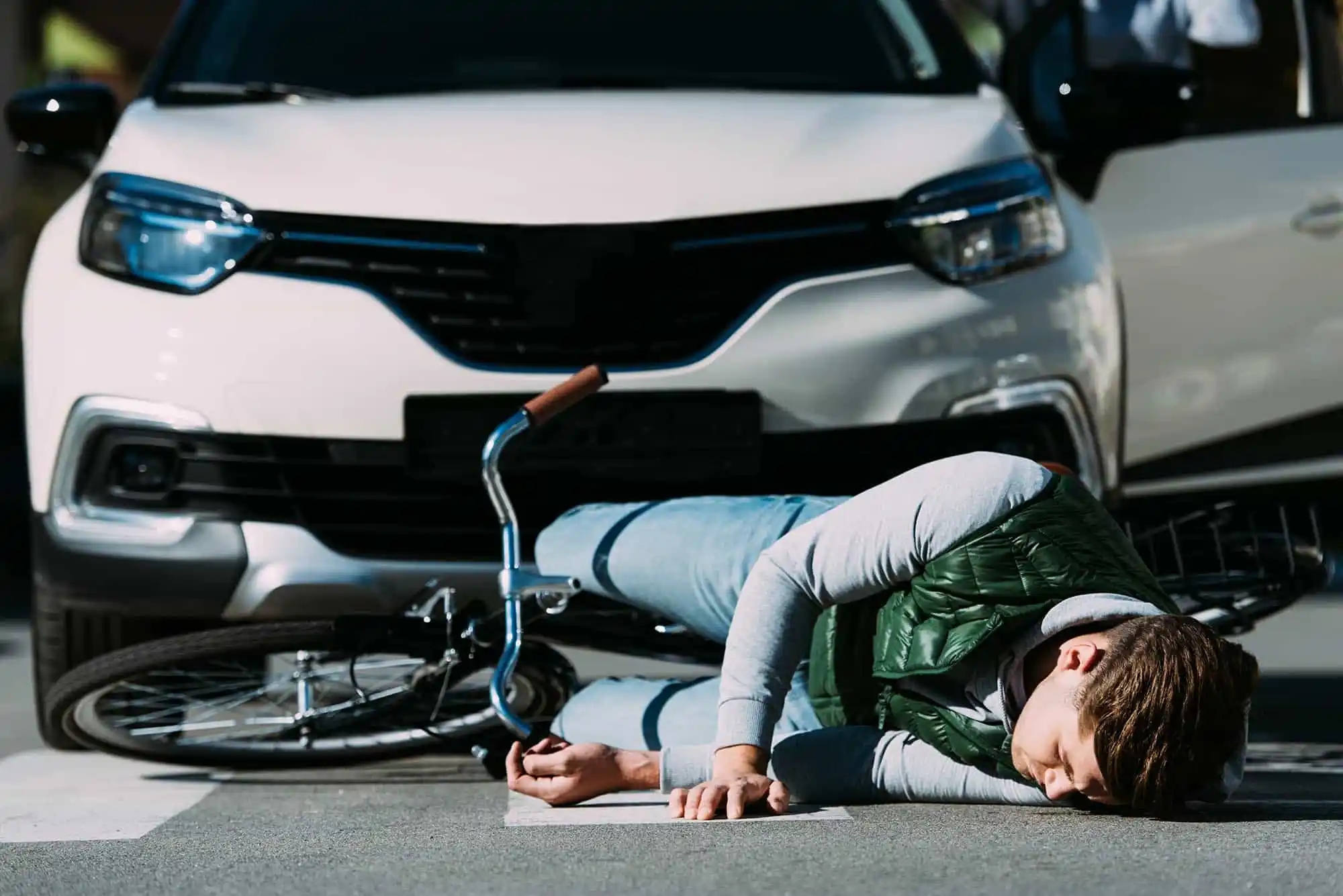 A person lies on the ground next to a fallen bicycle in front of a stopped white car, suggesting a collision at a crosswalk. - Ghassemi Law Group A person lies on the ground next to a fallen bicycle in front of a stopped white car, suggesting a collision at a crosswalk
