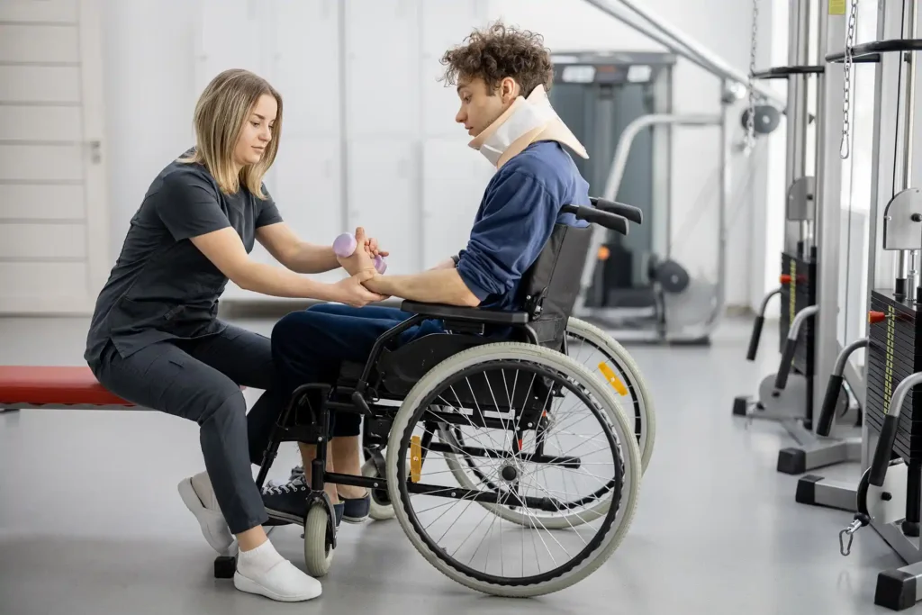 A physical therapist assists a man in a wheelchair wearing a neck brace with hand exercises in a rehabilitation facility