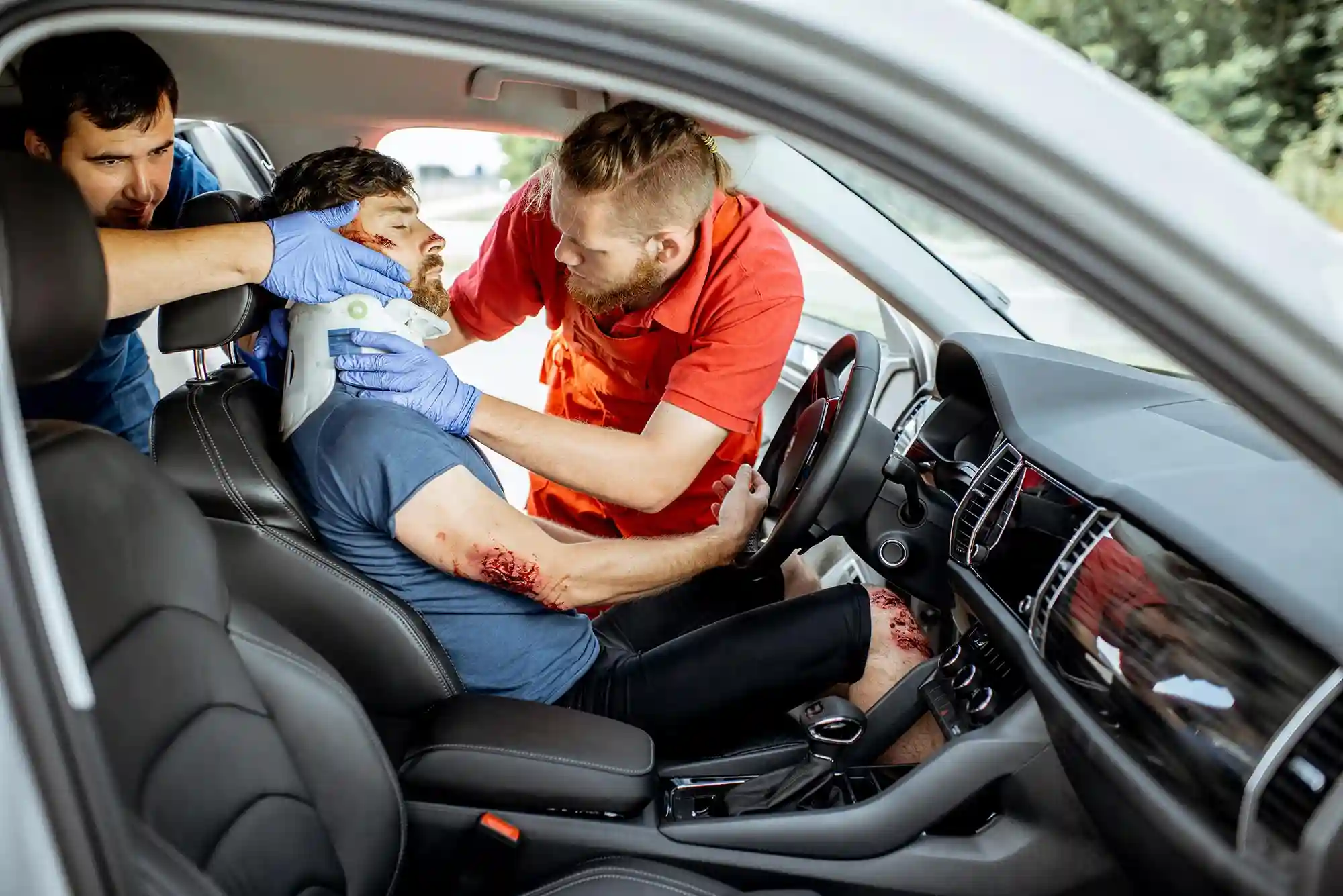 Two emergency responders fit a neck brace on an injured man in the driver’s seat of a car  The man has visible wounds on his arm and head  The car door is open 