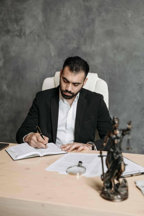 A man in a suit sits at a desk writing in a notebook, with legal documents, a magnifying glass, and a Lady Justice statue in front of him. - Ghassemi Law Group A man in a suit sits at a desk writing in a notebook, with legal documents, a magnifying glass, and a Lady Justice statue in front of him