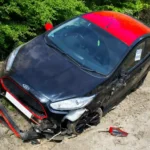 A black and red car, involved in an automobile accident, is off the road on a dirt area near green foliage  The car’s bumper is detached and a side mirror is broken, indicating significant front end damage 