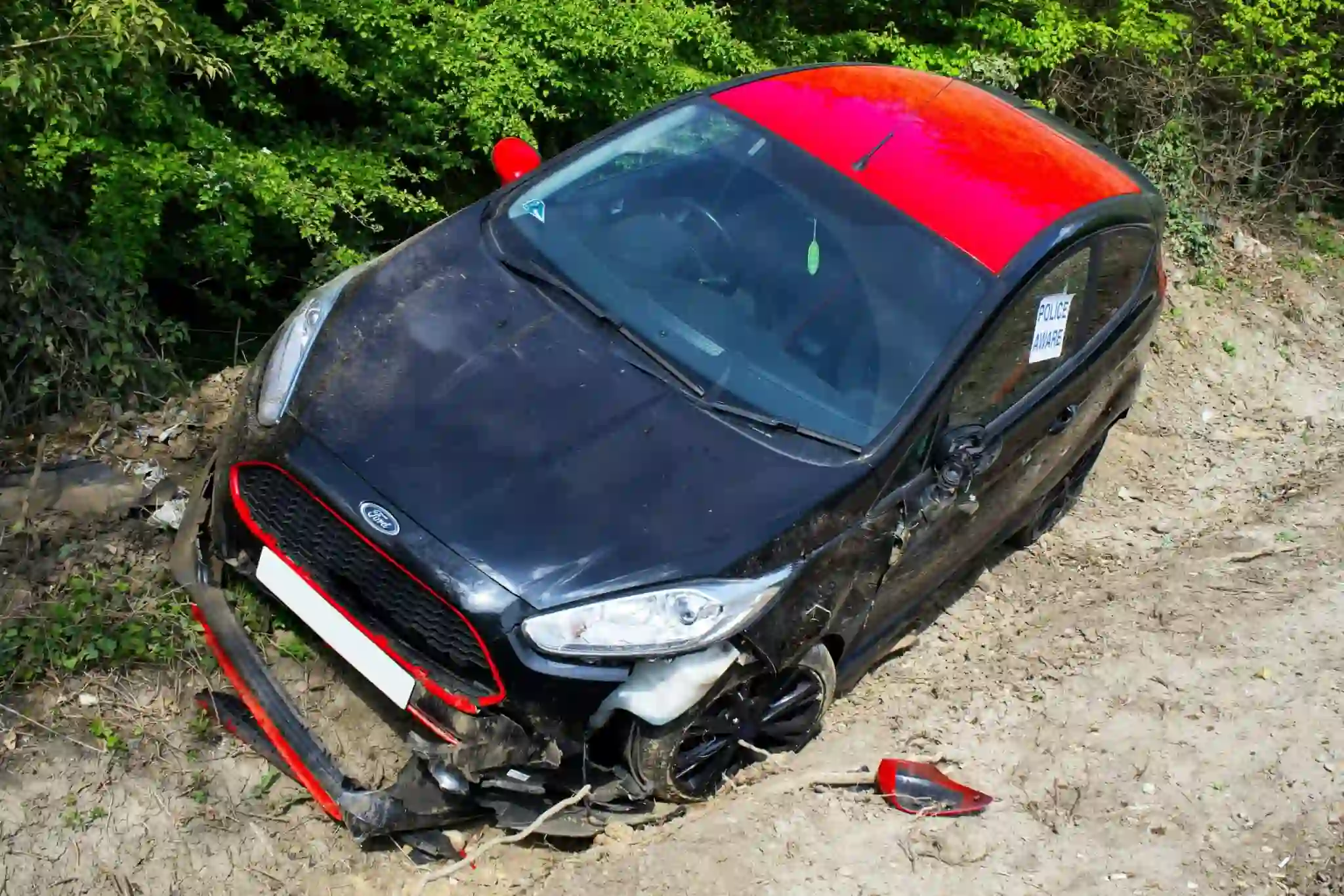 A black and red car, involved in an automobile accident, is off the road on a dirt area near green foliage  The car’s bumper is detached and a side mirror is broken, indicating significant front end damage 