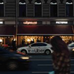A blurred figure crosses a city street in front of a white Uber car parked outside a restaurant with neon signs on a dark building, capturing the kind of moment that might require the expertise of an Uber Accident Lawyer