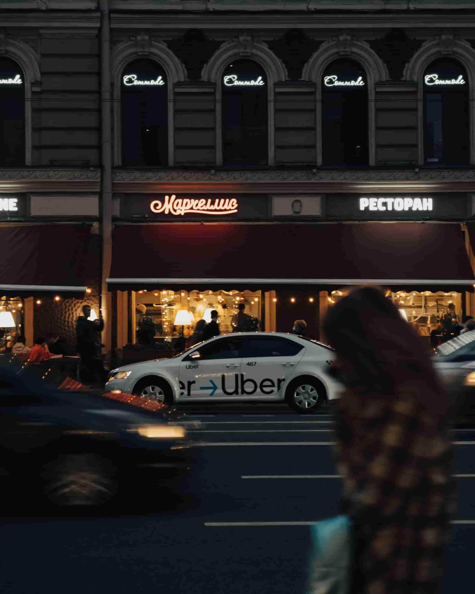 A blurred figure crosses a city street in front of a white Uber car parked outside a restaurant with neon signs on a dark building, capturing the kind of moment that might require the expertise of an Uber Accident Lawyer. - Ghassemi Law Group A blurred figure crosses a city street in front of a white Uber car parked outside a restaurant with neon signs on a dark building, capturing the kind of moment that might require the expertise of an Uber Accident Lawyer