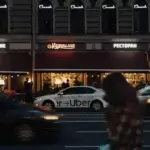 A blurred figure crosses a city street in front of a white Uber car parked outside a restaurant with neon signs on a dark building, capturing the kind of moment that might require the expertise of an Uber Accident Lawyer 
