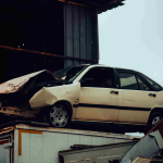 A damaged white car with a crumpled front end sits on top of debris near a metal building, showing the aftermath of a car accident. - Ghassemi Law Group A damaged white car with a crumpled front end sits on top of debris near a metal building, showing the aftermath of a car accident