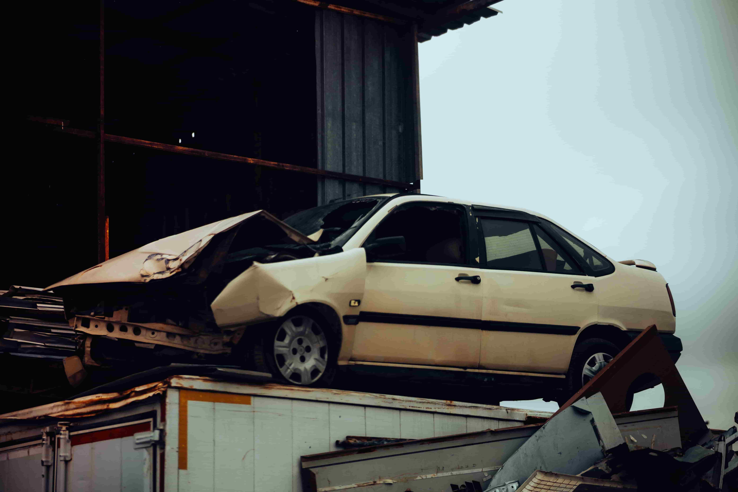 A damaged white car with a crumpled front end sits on top of debris near a metal building, showing the aftermath of a car accident. - Ghassemi Law Group A damaged white car with a crumpled front end sits on top of debris near a metal building, showing the aftermath of a car accident