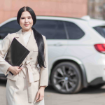 Woman in a light colored coat stands outdoors holding a black folder, with a white SUV parked in the background. - Ghassemi Law Group Woman in a light colored coat stands outdoors holding a black folder, with a white SUV parked in the background