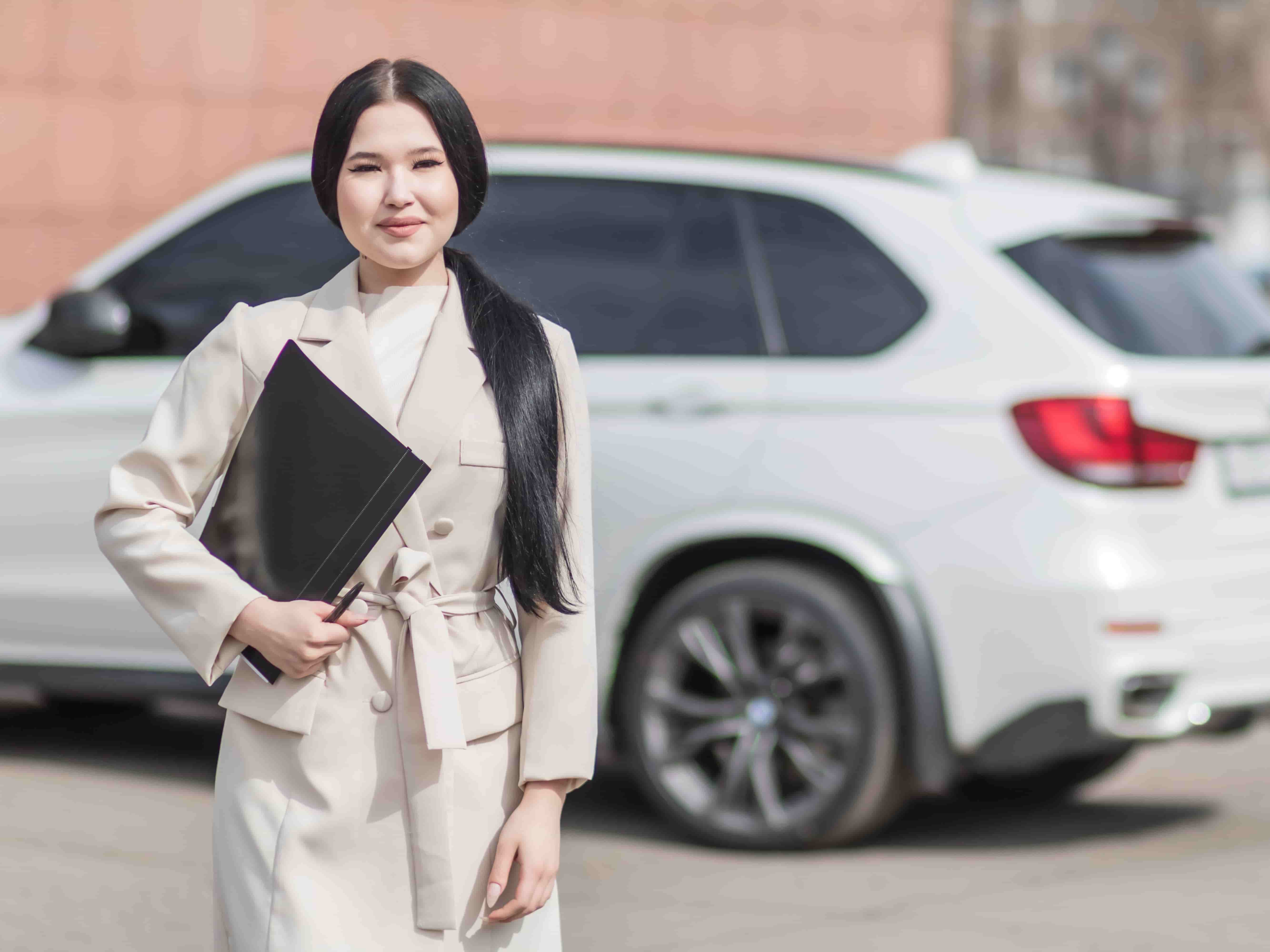 Woman in a light colored coat stands outdoors holding a black folder, with a white SUV parked in the background. - Ghassemi Law Group Woman in a light colored coat stands outdoors holding a black folder, with a white SUV parked in the background
