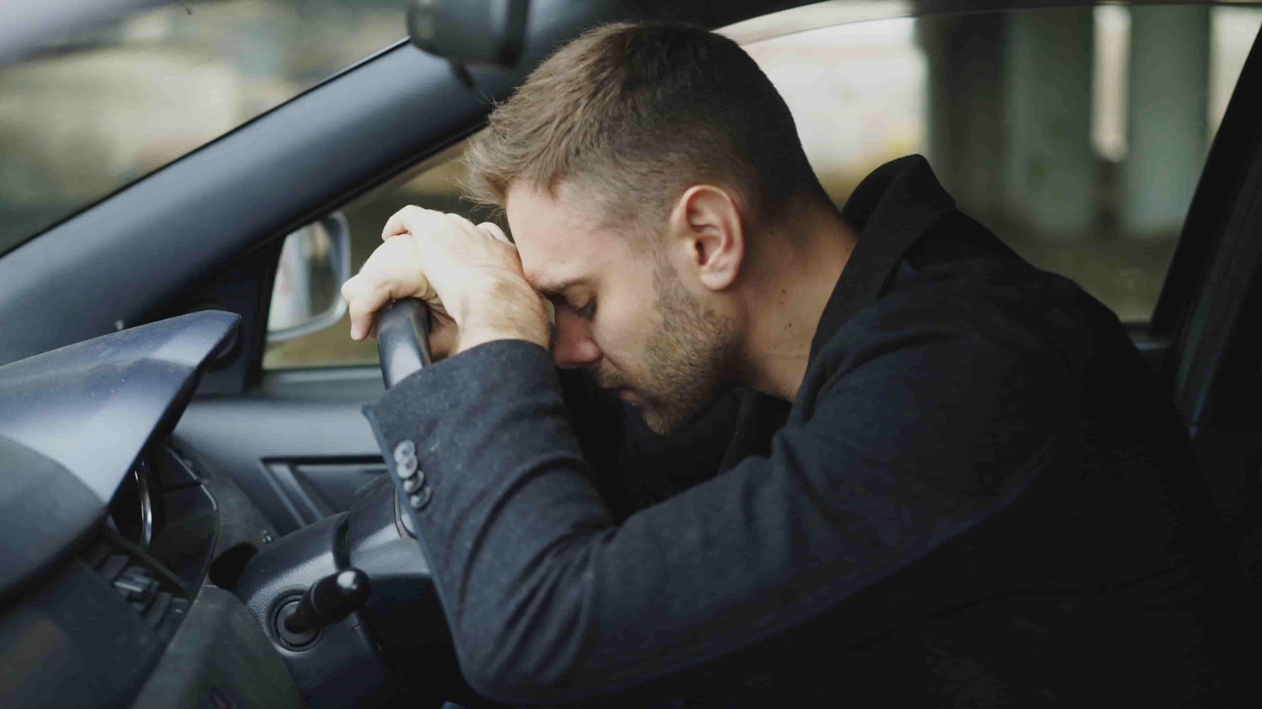 A man sits in the driver’s seat of a car, resting his forehead on the steering wheel with his eyes closed, appearing stressed or exhausted—perhaps wondering, "How long will it take to settle my car accident case in Irvine?. - Ghassemi Law Group A man sits in the driver’s seat of a car, resting his forehead on the steering wheel with his eyes closed, appearing stressed or exhausted—perhaps wondering, "How long will it take to settle my car accident case in Irvine?