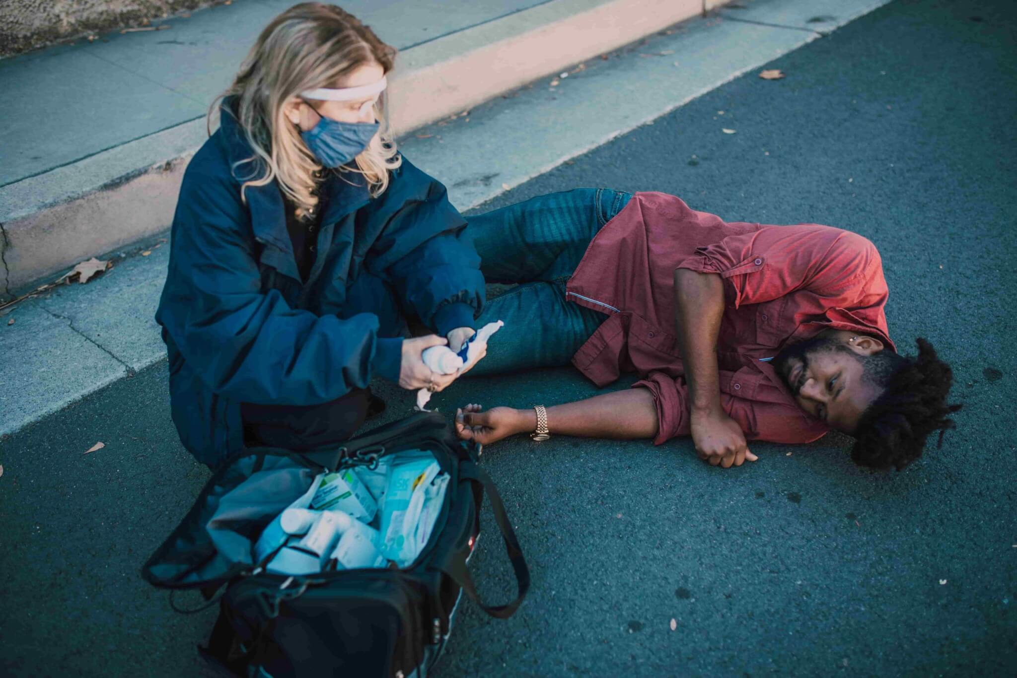 A person wearing a mask and gloves prepares medical supplies next to a man lying on his side on the pavement near a curb, raising questions such as, "How long do I have to file a pedestrian accident claim in California?. - Ghassemi Law Group A person wearing a mask and gloves prepares medical supplies next to a man lying on his side on the pavement near a curb, raising questions such as, "How long do I have to file a pedestrian accident claim in California?