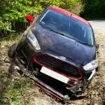 A black and red car was involved in a car accident, veering off the road into a ditch with significant front end damage and a detached front bumper