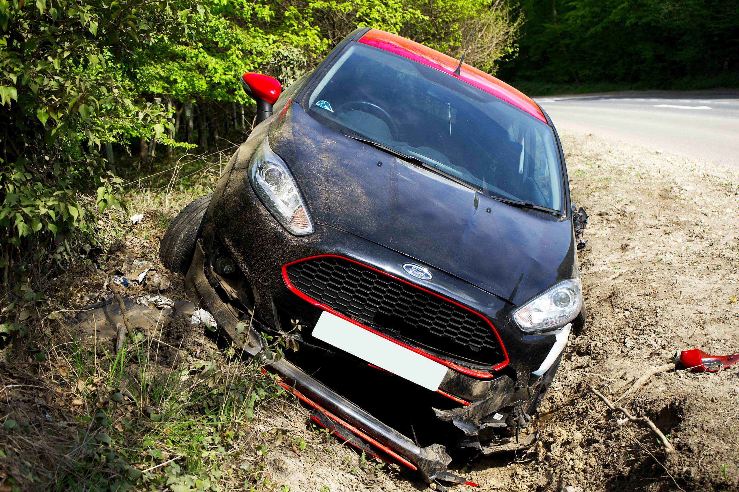 A black and red car was involved in a car accident, veering off the road into a ditch with significant front end damage and a detached front bumper. - Ghassemi Law Group A black and red car was involved in a car accident, veering off the road into a ditch with significant front end damage and a detached front bumper