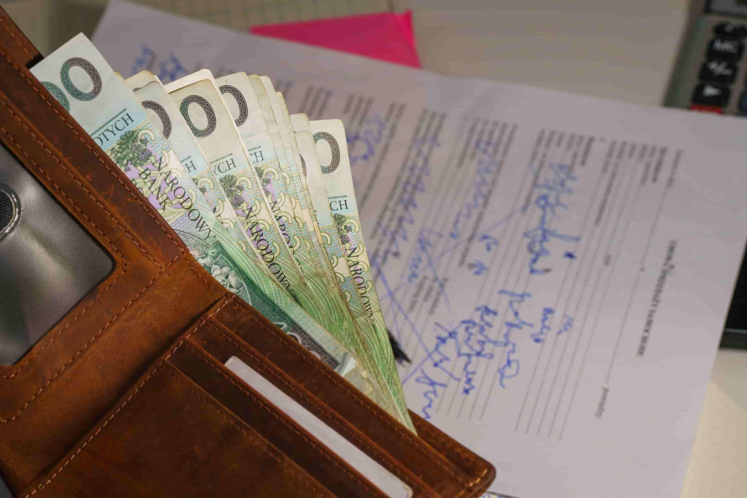 A brown wallet filled with Polish banknotes is placed on top of a handwritten document with blue ink, resembling files you might see when researching What Is the Average Settlement for a Car Accident Injury in Irvine. - Ghassemi Law Group A brown wallet filled with Polish banknotes is placed on top of a handwritten document with blue ink, resembling files you might see when researching What Is the Average Settlement for a Car Accident Injury in Irvine