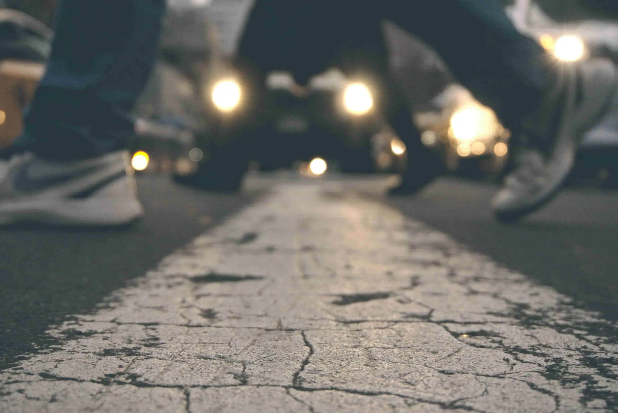 Close up of a cracked street line with two people crossing, their legs in motion, and blurred car headlights in the background. - Ghassemi Law Group Close up of a cracked street line with two people crossing, their legs in motion, and blurred car headlights in the background