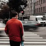A person in a red jacket stands on a crosswalk, facing traffic with blurred vehicles passing by in an urban setting. - Ghassemi Law Group A person in a red jacket stands on a crosswalk, facing traffic with blurred vehicles passing by in an urban setting