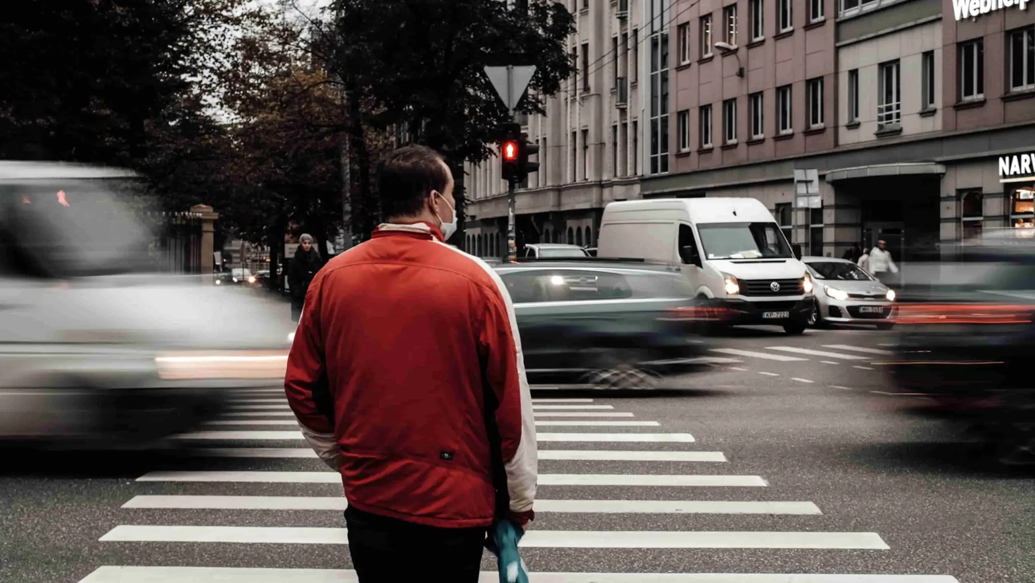 A person in a red jacket stands on a crosswalk, facing traffic with blurred vehicles passing by in an urban setting. - Ghassemi Law Group A person in a red jacket stands on a crosswalk, facing traffic with blurred vehicles passing by in an urban setting