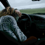 Person wearing a patterned sweater sits slouched with their head resting on the steering wheel inside a car on a rainy day, looking out at a foggy field. - Ghassemi Law Group Person wearing a patterned sweater sits slouched with their head resting on the steering wheel inside a car on a rainy day, looking out at a foggy field