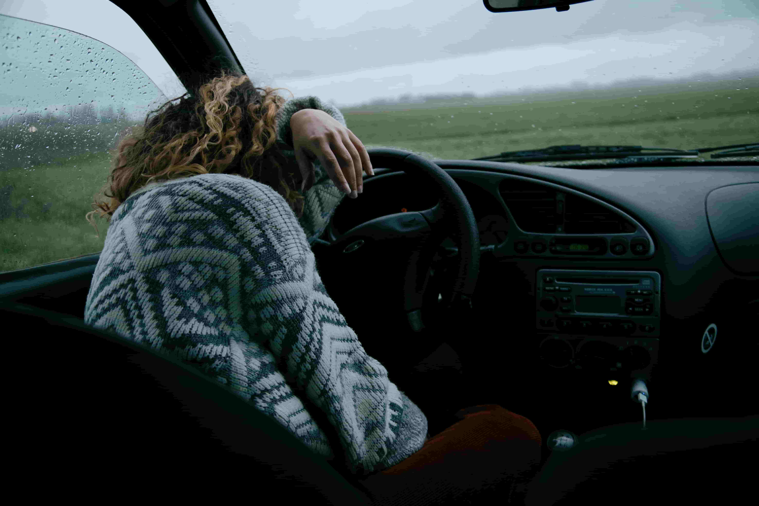 Person wearing a patterned sweater sits slouched with their head resting on the steering wheel inside a car on a rainy day, looking out at a foggy field. - Ghassemi Law Group Person wearing a patterned sweater sits slouched with their head resting on the steering wheel inside a car on a rainy day, looking out at a foggy field