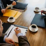 Three people sit at a wooden table with folders, notepads, and coffee cups, engaged in a business meeting or discussion  One person is writing in a notebook 