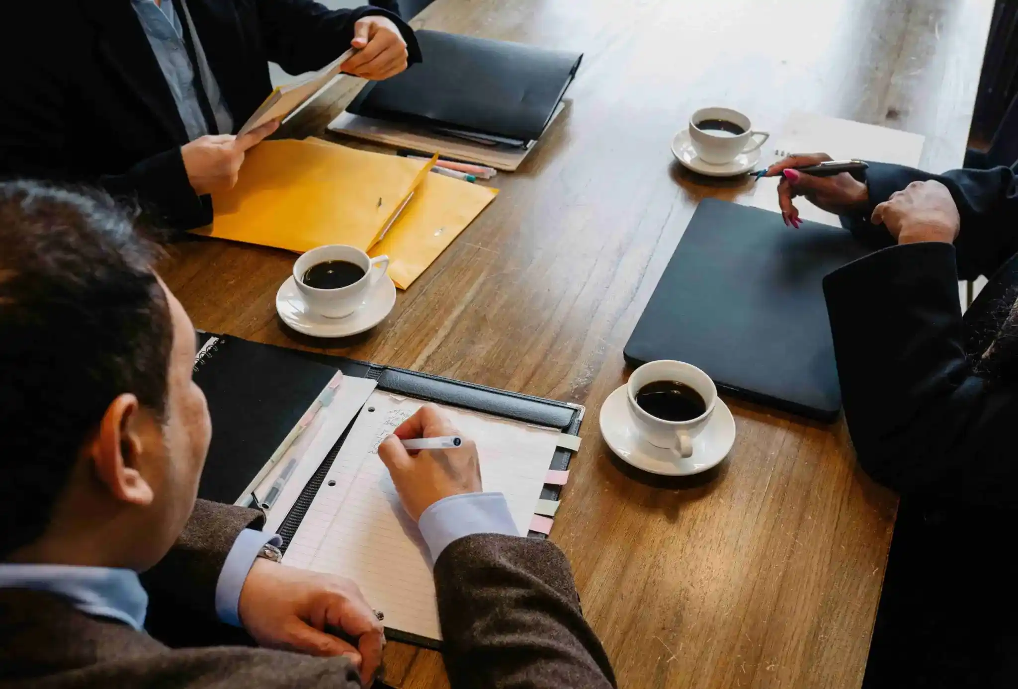 Three people sit at a wooden table with folders, notepads, and coffee cups, engaged in a business meeting or discussion  One person is writing in a notebook 